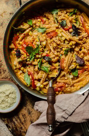 An overhead shot shows a creamy red pepper pasta in a stainless steel pan. The short-shaped pasta is garnished with fresh basil leaves, strips of roasted red pepper, charred corn kernels, dried chili flakes, and freshly ground pepper.