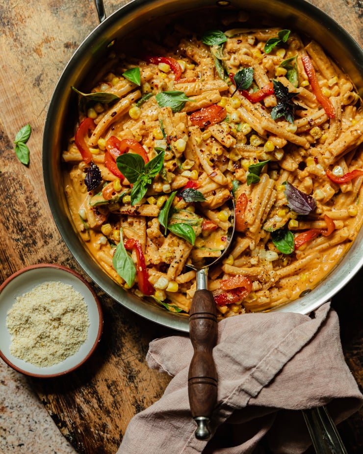 An overhead shot shows a creamy red pepper pasta in a stainless steel pan. The short-shaped pasta is garnished with fresh basil leaves, strips of roasted red pepper, charred corn kernels, dried chili flakes, and freshly ground pepper.