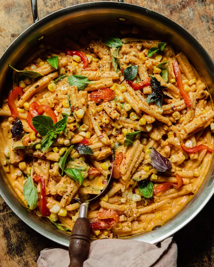 An overhead shot shows a creamy vegan red pepper pasta in a stainless steel pan. The short-shaped pasta is garnished with fresh basil leaves, strips of roasted red pepper, charred corn kernels, dried chili flakes, and freshly ground pepper.