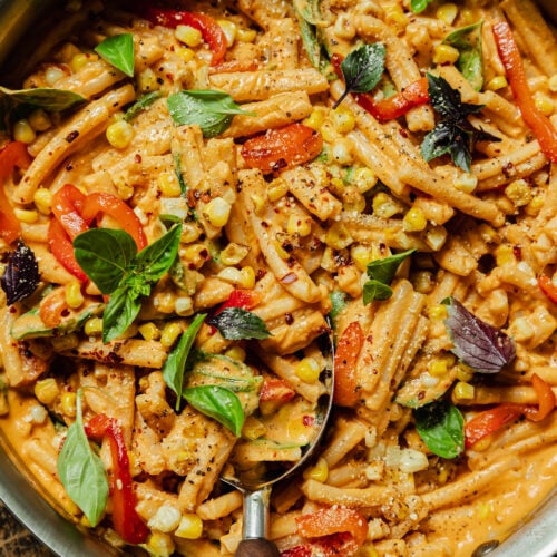 An up close, overhead shot shows a creamy red pepper pasta in a stainless steel pan. The short-shaped pasta is garnished with fresh basil leaves, strips of roasted red pepper, charred corn kernels, dried chili flakes, and freshly ground pepper.