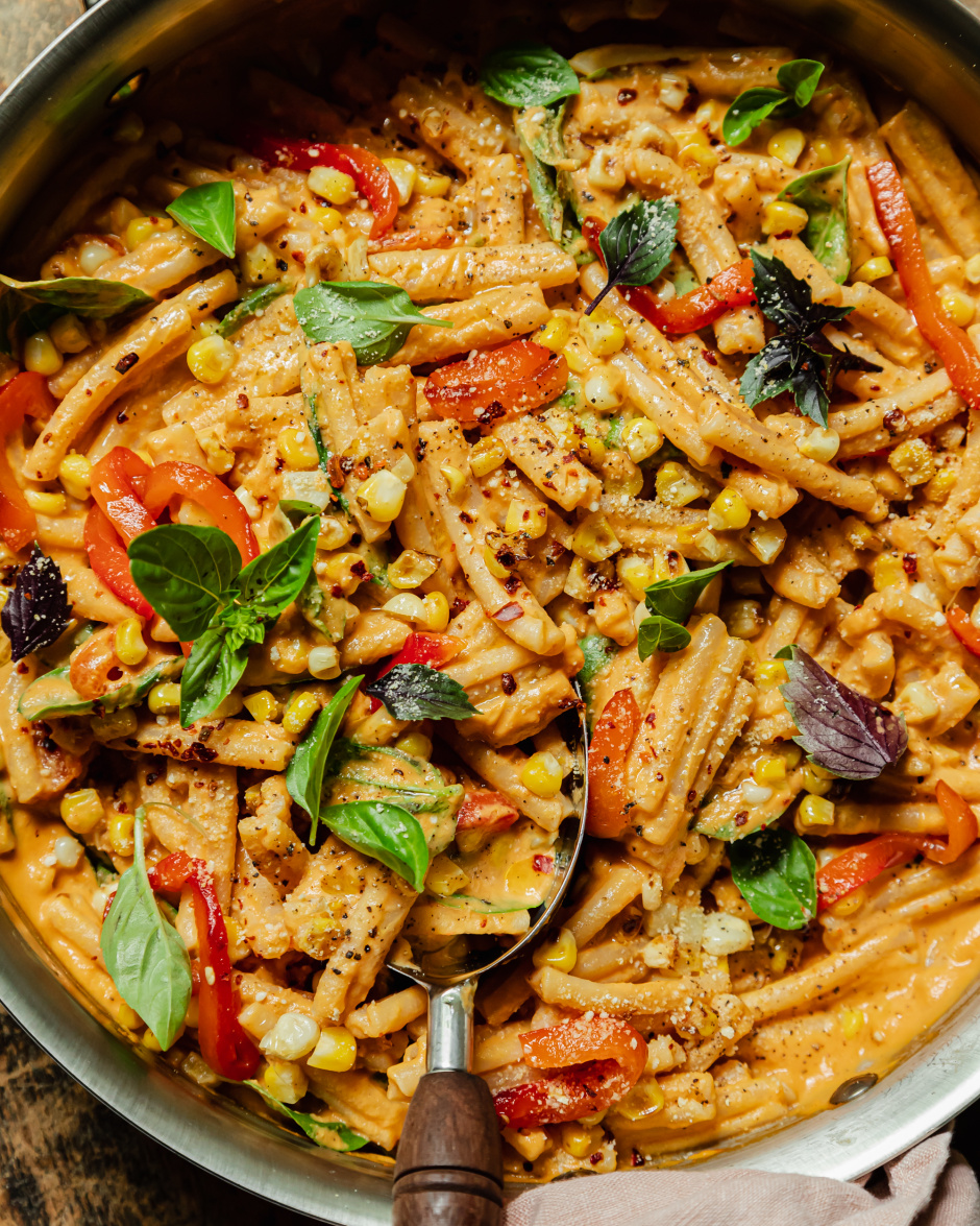 An up close, overhead shot shows a creamy red pepper pasta in a stainless steel pan. The short-shaped pasta is garnished with fresh basil leaves, strips of roasted red pepper, charred corn kernels, dried chili flakes, and freshly ground pepper.
