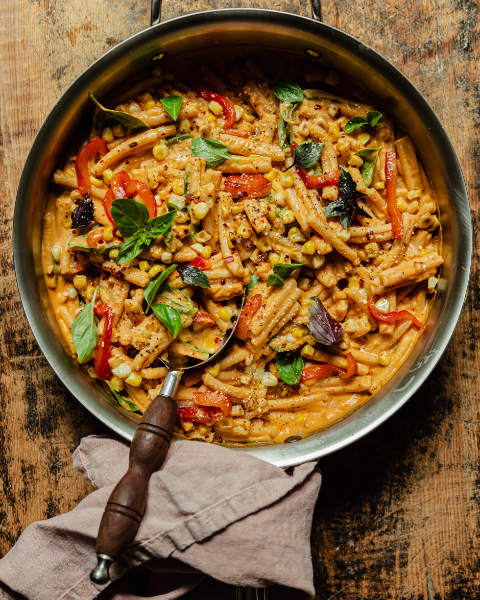 An overhead shot shows a creamy vegan red pepper pasta in a stainless steel pan. The short-shaped pasta is garnished with fresh basil leaves, strips of roasted red pepper, charred corn kernels, dried chili flakes, and freshly ground pepper.