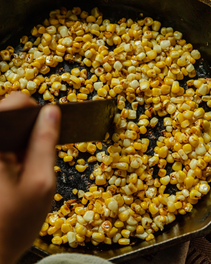 An overhead image shows a hand stirring some charred corn with a wooden utensil in a cast iron skillet.