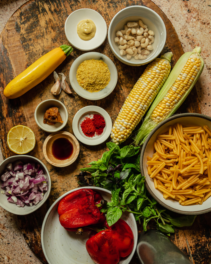 An overhead shot of ingredients used in a vegan red pepper pasta dish that also features yellow zucchini, corn, and fresh basil. All ingredients are on a vintage, rustic wood board.