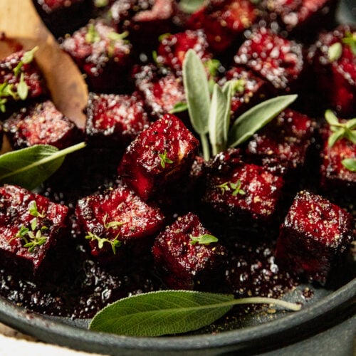 An up close, 3/4 angle shot of a sauté pan filled with blackberry glazed tofu cubes. The glaze is deep fuchsia and the dish is garnished with fresh sage and thyme leaves.