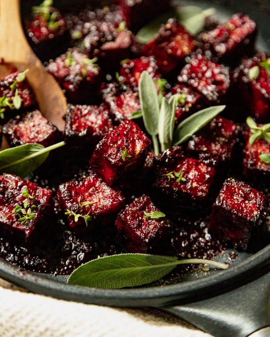 An up close, 3/4 angle shot of a sauté pan filled with blackberry glazed tofu cubes. The glaze is deep fuchsia and the dish is garnished with fresh sage and thyme leaves.