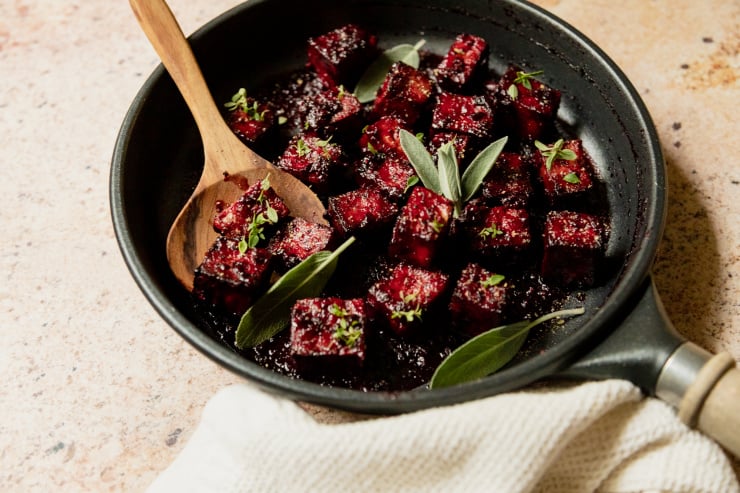 A slight 3/4 angle shot of a sauté pan filled with blackberry glazed tofu cubes. The glaze is deep fuchsia and the dish is garnished with fresh sage and thyme leaves.