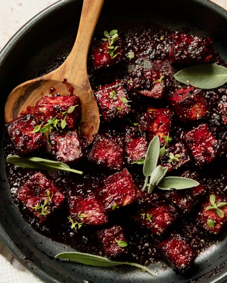 An up close, overhead shot of a sauté pan filled with blackberry glazed tofu cubes. The glaze is deep fuchsia and the dish is garnished with fresh sage and thyme leaves.