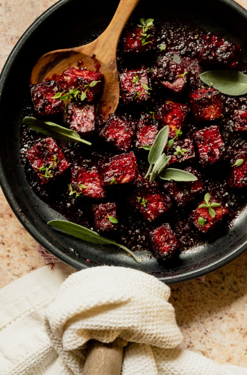 An overhead shot of a sauté pan filled with blackberry glazed tofu cubes. The glaze is deep fuchsia and the dish is garnished with fresh sage and thyme leaves.