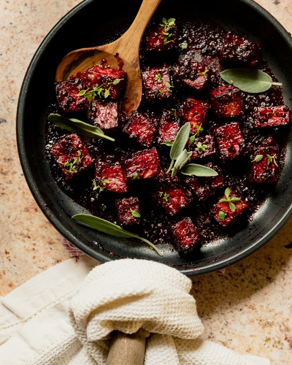 An overhead shot of a sauté pan filled with blackberry glazed tofu cubes. The glaze is deep fuchsia and the dish is garnished with fresh sage and thyme leaves.