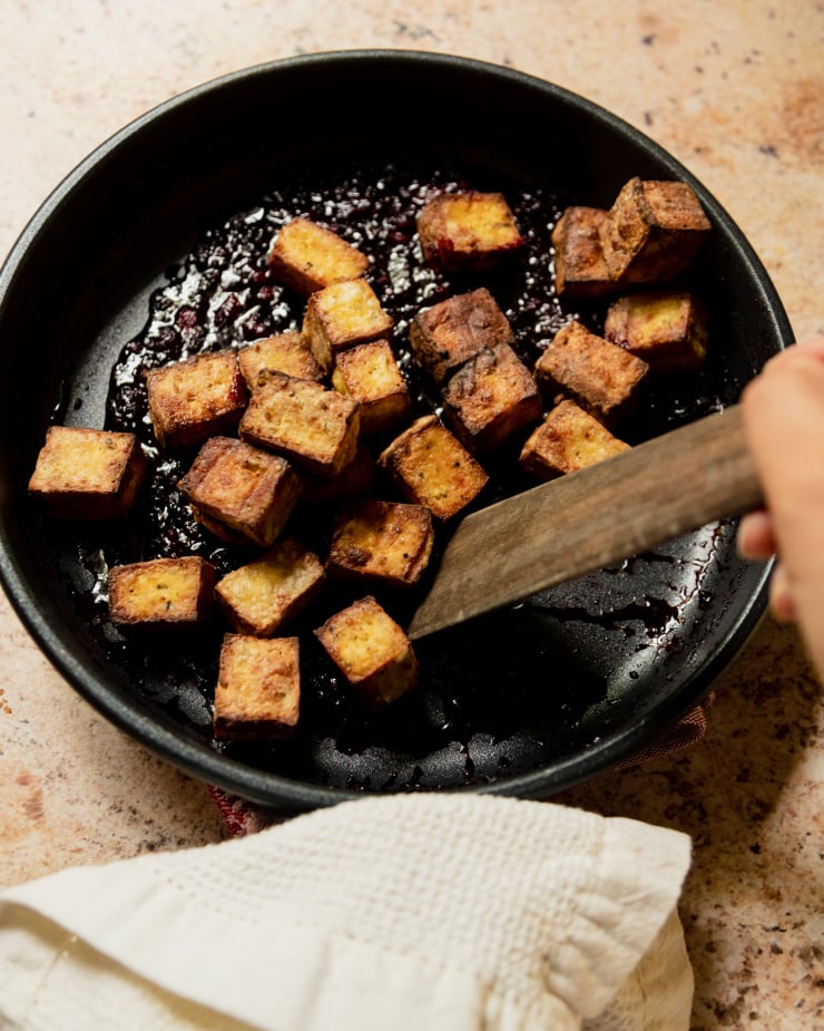 Image shows a hand using a wooden utensil to stir crispy cubes of tofu in a blackberry glaze.