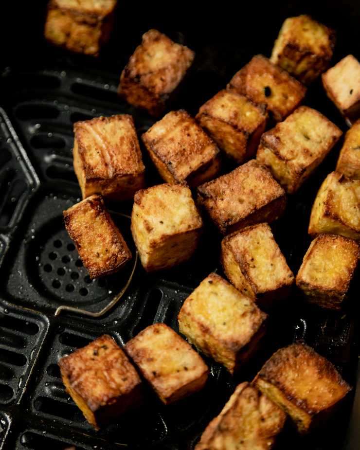 A 3/4 angle shot shows crispy and golden cubes of tofu inside of a dark air fryer basket.