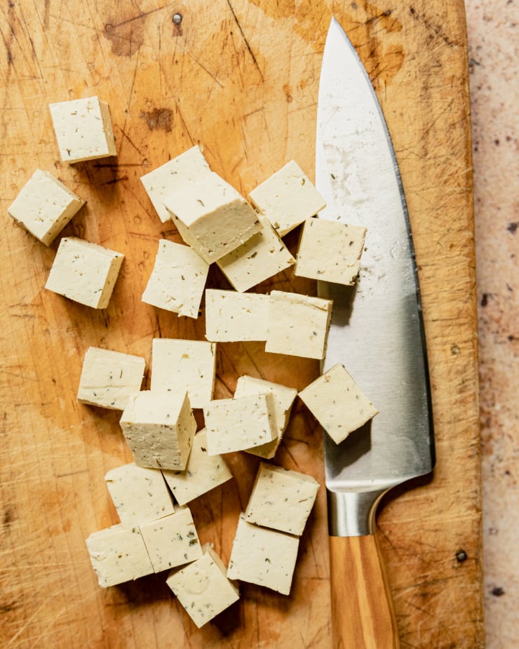 An overhead shot shows cubes of raw tofu on a wooden cutting board next to a chef's knife.