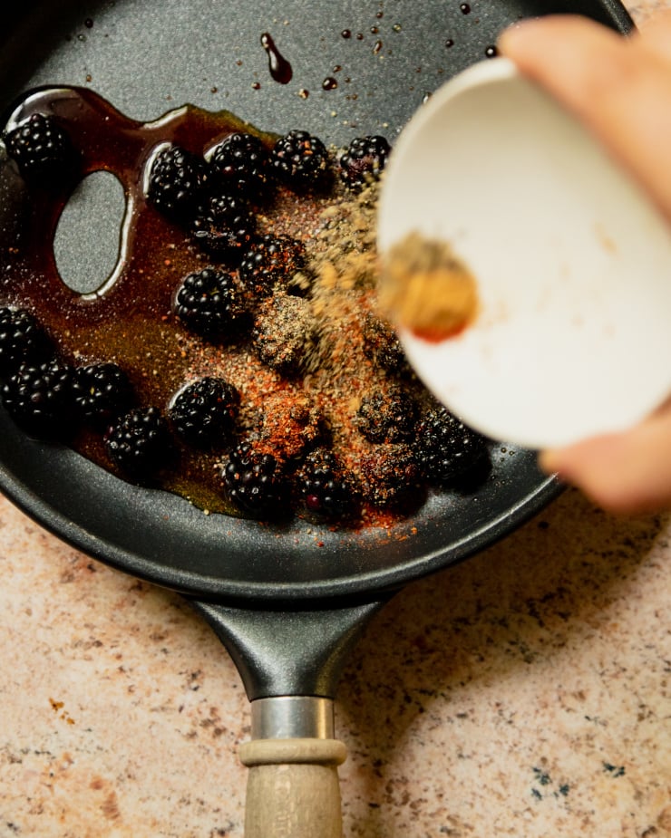 An overhead shot shows a hand emptying spices from a white bowl into a sauté pan of whole fresh blackberries.