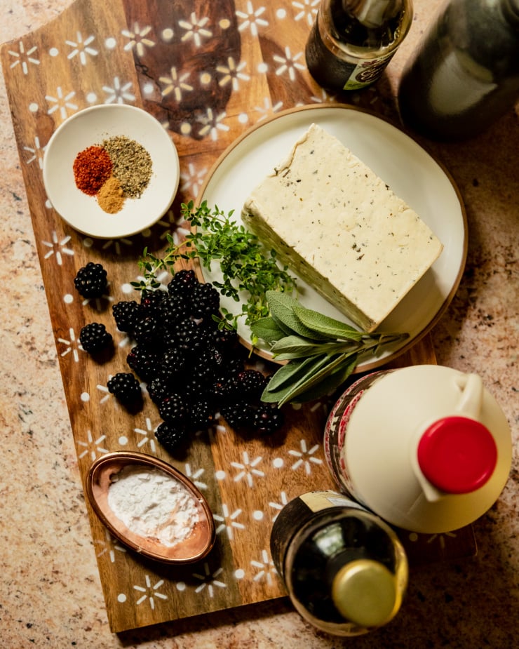 An overhead shot of ingredients for a blackberry tofu recipe. All ingredients are on an etched wooden cutting board on a pink/tan stone background.