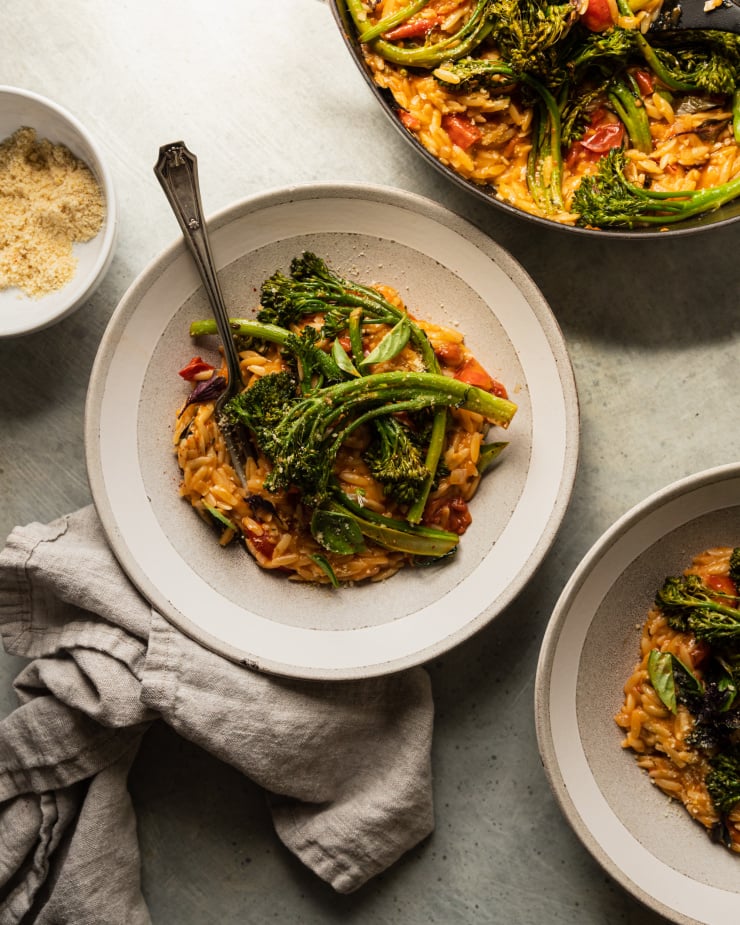 An overhead shot of a vegan tomato orzo dish, served up in a light grey serving bowl. The orzo is topped with roasted broccolini.