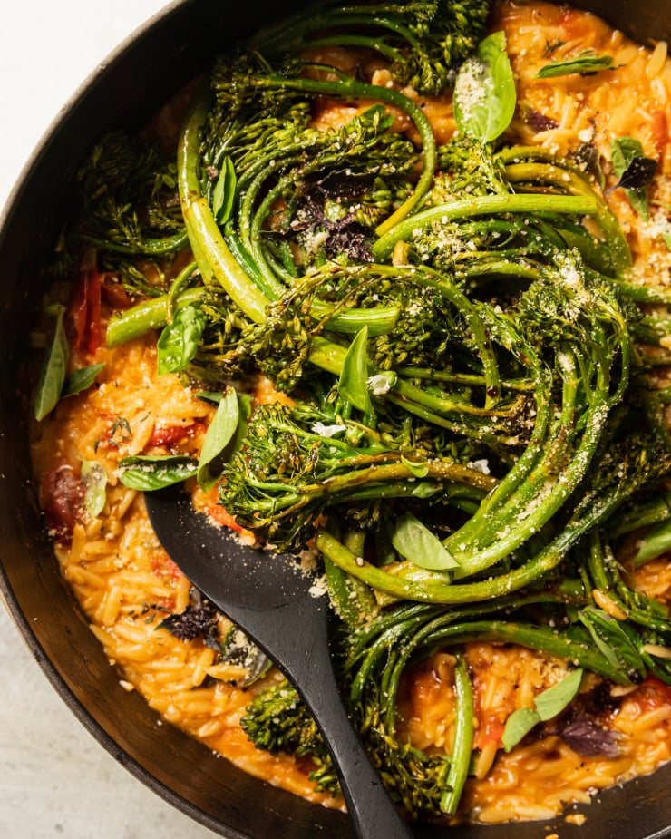 An up close overhead shot of a creamy vegan tomato orzo dish in a pot with roasted broccolini and fresh basil on top.
