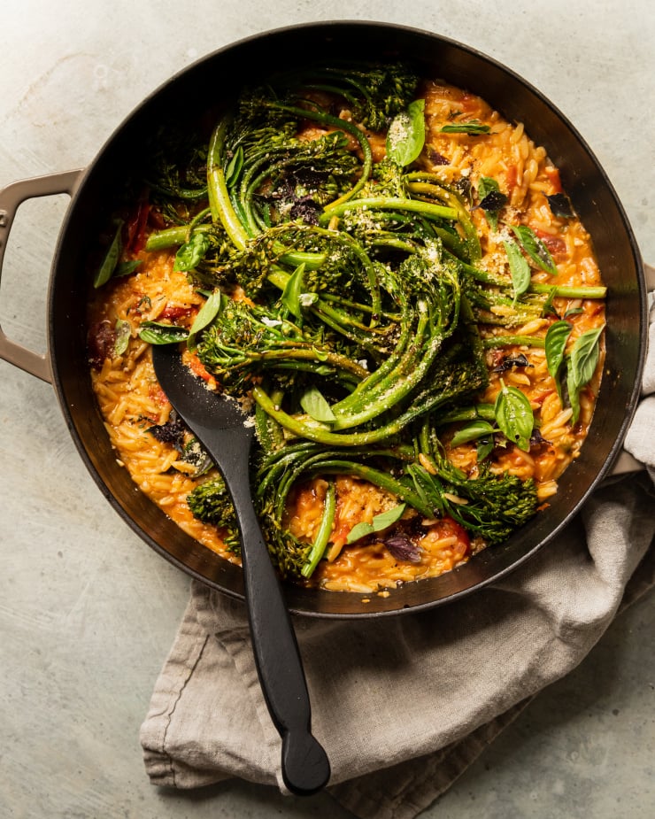 An overhead shot of a creamy vegan tomato orzo dish in a pot with roasted broccolini and fresh basil on top.
