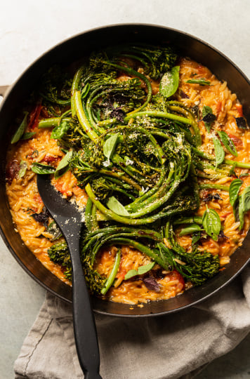 An overhead shot of a creamy vegan tomato orzo dish in a pot with roasted broccolini and fresh basil on top.