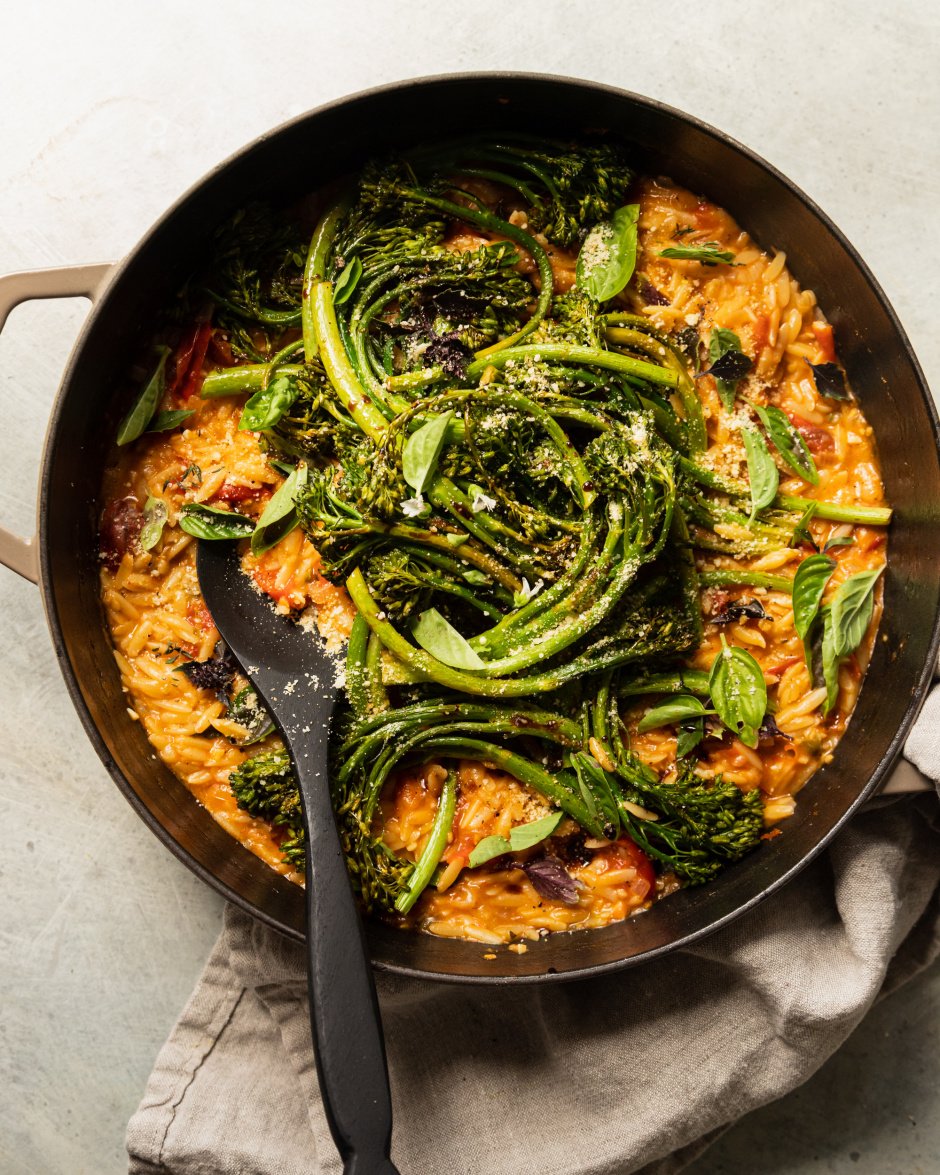 An overhead shot of a creamy vegan tomato orzo dish in a pot with roasted broccolini and fresh basil on top.