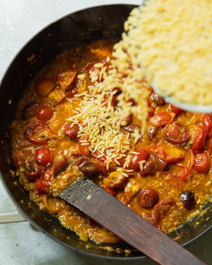 An overhead shot shows dry orzo pasta being poured into a pot with sautéed tomatoes.