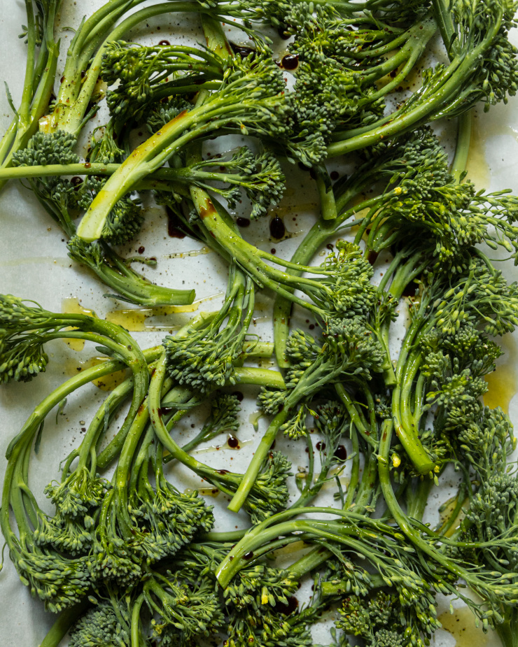 An up close, overhead shot shows broccolini stalks on a parchment lined baking sheet, drizzled with olive oil and balsamic vinegar and seasoned with salt and pepper.