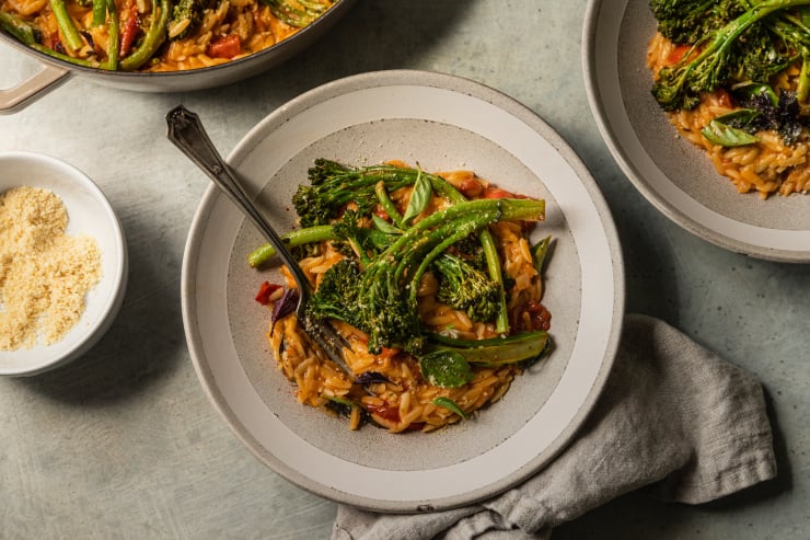 An overhead shot of a vegan tomato orzo dish, served up in a light grey serving bowl. The orzo is topped with roasted broccolini.