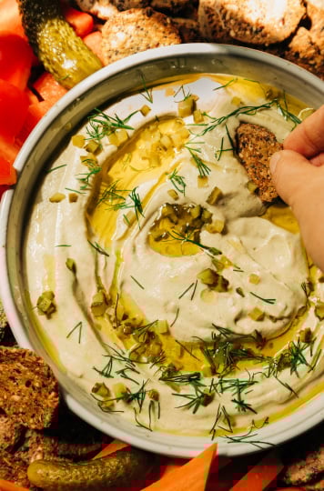 An overhead image shows a bowl of a creamy, beige vegan dill pickle dip that's swirled with olive oil on the top and garnished with fresh dill and finely chopped pickles. The dip is served with crackers and cut vegetables. A hand is seen dragging a cracker through the dip.