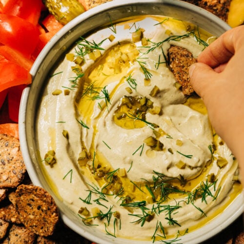 An overhead image shows a bowl of a creamy, beige vegan dill pickle dip that's swirled with olive oil on the top and garnished with fresh dill and finely chopped pickles. The dip is served with crackers and cut vegetables. A hand is seen dragging a cracker through the dip.