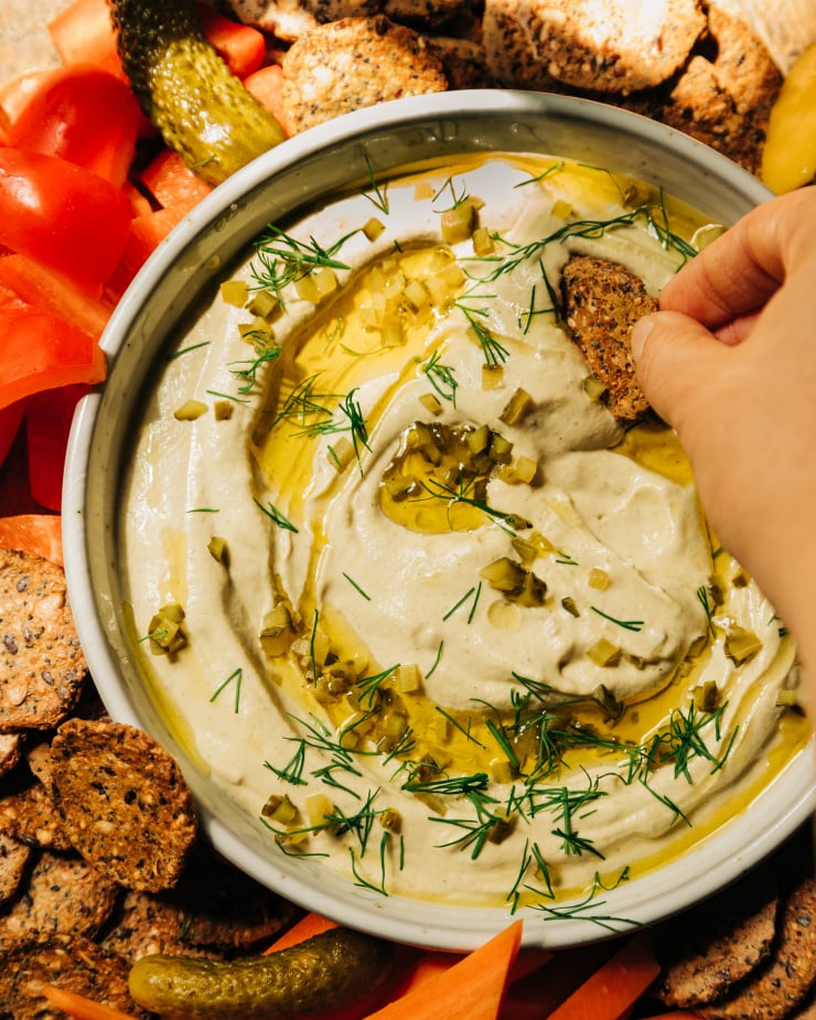 An overhead image shows a bowl of a creamy, beige vegan dill pickle dip that's swirled with olive oil on the top and garnished with fresh dill and finely chopped pickles. The dip is served with crackers and cut vegetables. A hand is seen dragging a cracker through the dip.
