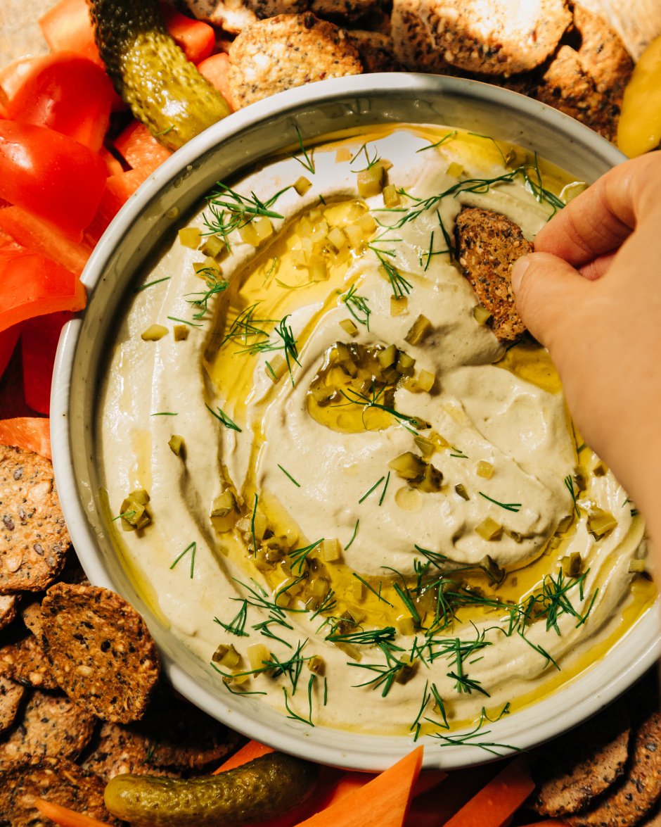 An overhead image shows a bowl of a creamy, beige vegan dill pickle dip that's swirled with olive oil on the top and garnished with fresh dill and finely chopped pickles. The dip is served with crackers and cut vegetables. A hand is seen dragging a cracker through the dip.