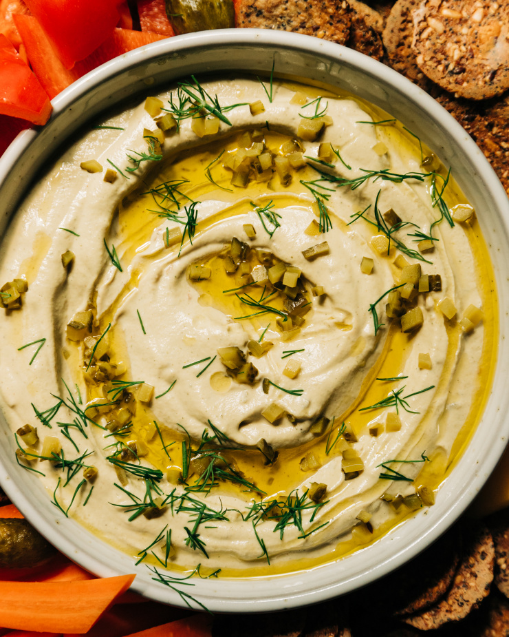 An overhead image shows a bowl of a creamy, beige vegan dill pickle dip that's swirled with olive oil on the top and garnished with fresh dill and finely chopped pickles. The dip is served with crackers and cut vegetables.