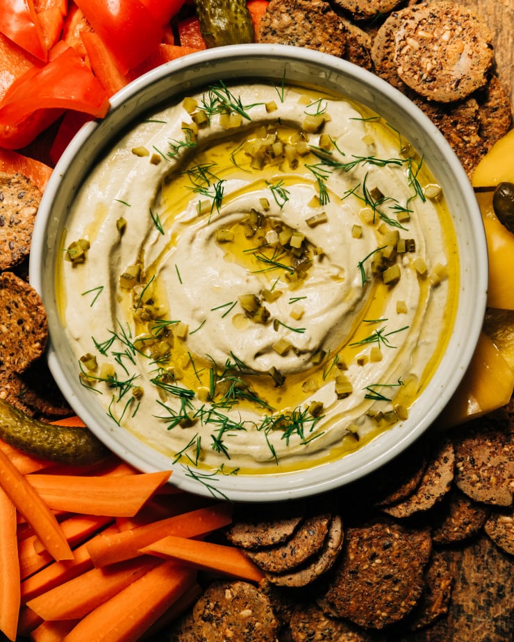 An overhead image shows a bowl of a creamy, beige vegan dill pickle dip that's swirled with olive oil on the top and garnished with fresh dill and finely chopped pickles. The dip is served with crackers and cut vegetables.