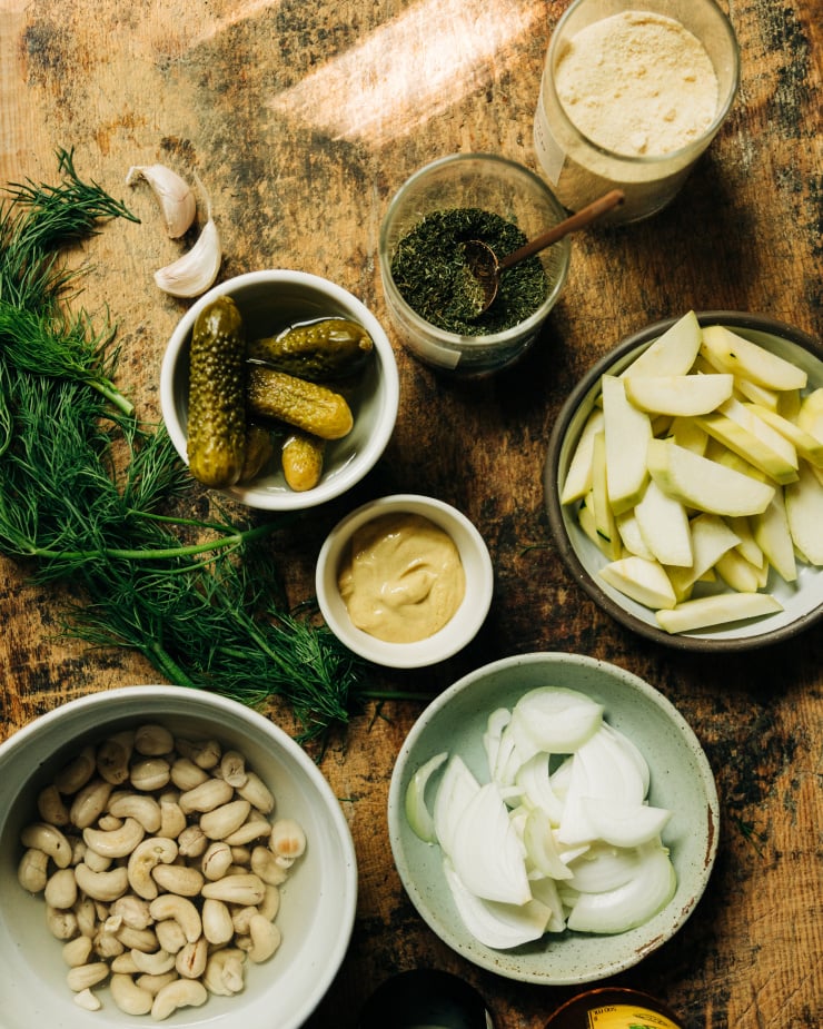 An overhead image shows ingredients on a rough wooden board, ready to be used in a vegan dill pickle dip.