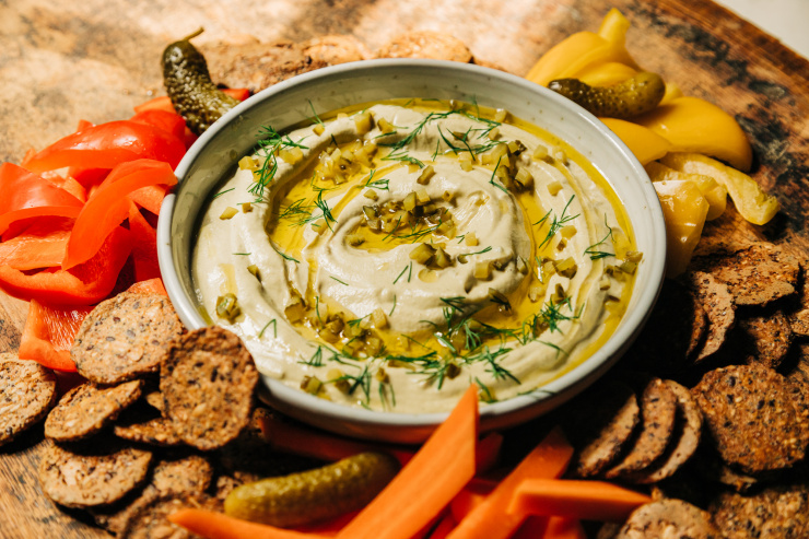 A 3/4 angle image shows a bowl of a creamy, beige vegan dill pickle dip that's swirled with olive oil on the top and garnished with fresh dill and finely chopped pickles. The dip is served with crackers and cut vegetables.