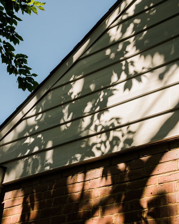 Image shows shadows from tree branches on the white siding of a house. Blue sky is in the background.