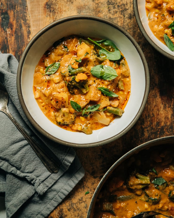An overhead shot of a creamy vegan vegetable stew in white ceramic bowls with dark grey rims. The stew is light orange and features baby spinach, fresh basil, and sun dried tomatoes.