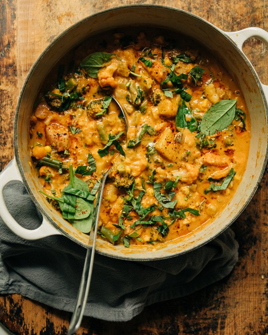 An overhead shot of a light orange creamy vegan vegetable stew that features sun dried tomatoes, baby spinach, and garlic. The stew is in a beige Dutch oven-style pot.