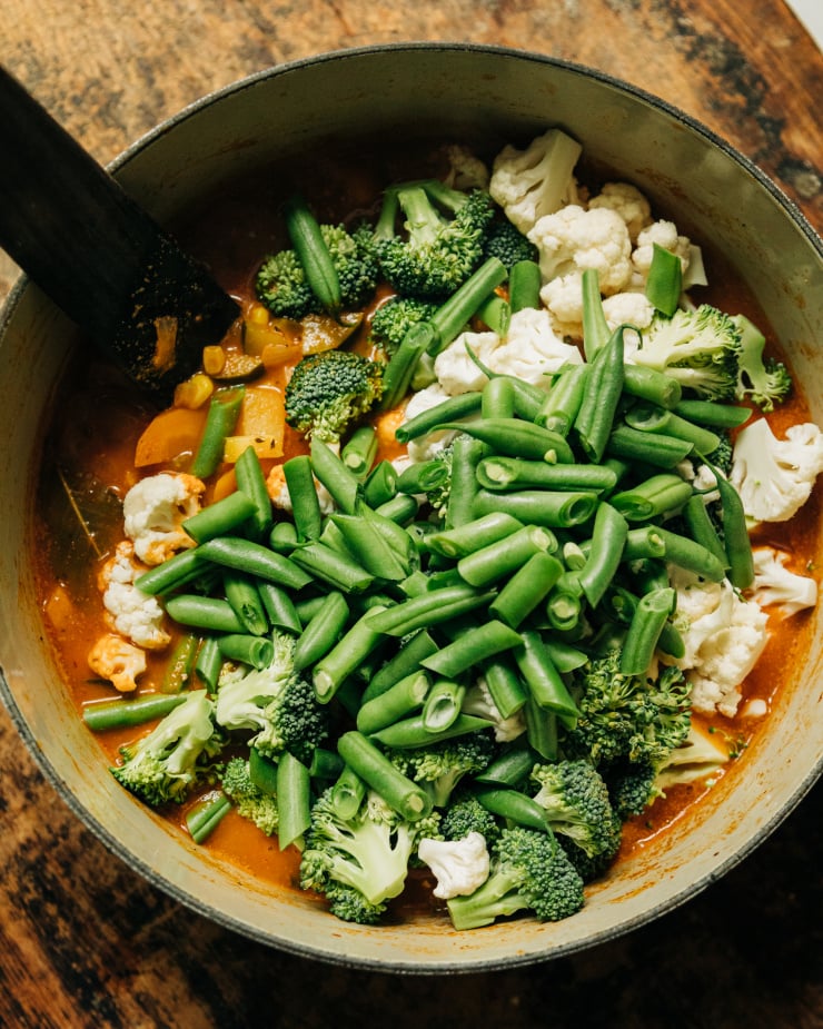 An overhead image shows cut green beans, broccoli, and cauliflower being added to a deep orange stew.