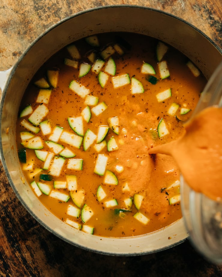 An overhead shot shows a deep orange, creamy sun dried tomato mixture being poured into a pot of stew.