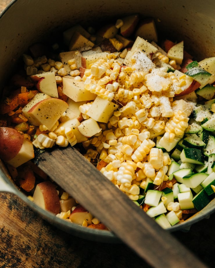 A slight 3/4 angle shot shows corn kernels, potatoes, and chopped zucchini being added to a pot of soup.