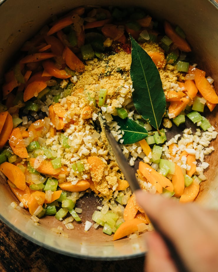 An overhead shot shows a wooden stirring utensil mixing up some aromatics in a soup pot: garlic, bay leaves, nutritional yeast, paprika, dried oregano, and thyme.