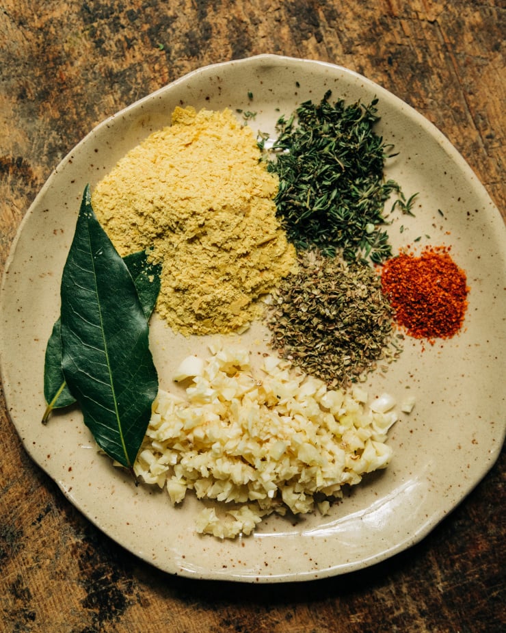 An overhead shot shows flavour enhancers used for a creamy vegan vegetable stew on a beige plate: bay leaves, minced garlic, ground chillies, dried oregano, and thyme.