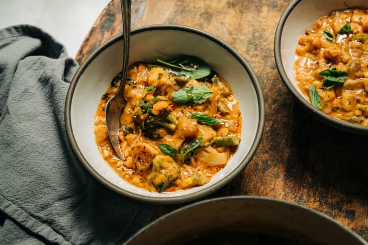 An overhead shot of a creamy vegan vegetable stew in white ceramic bowls with dark grey rims. The stew is light orange and features baby spinach, fresh basil, and sun dried tomatoes.