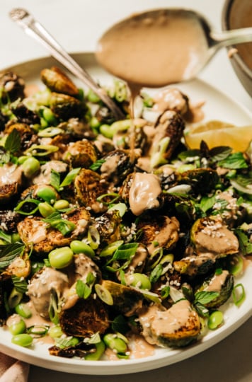 An up close, slight 3/4 angle shot of a plate of crispy roasted brussels sprouts and cooked edamame beans, drizzled with a creamy peanut sauce and finished with chopped green onions, mint, and sesame seeds.