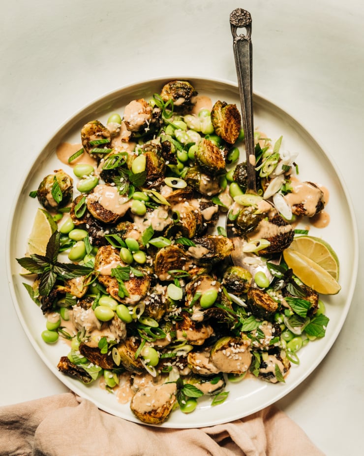 An overhead shot of a plate of crispy roasted brussels sprouts and cooked edamame beans, drizzled with a creamy peanut sauce and finished with chopped green onions, mint, and sesame seeds.