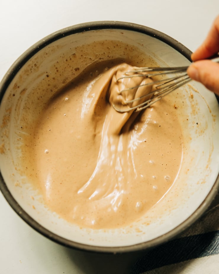 An overhead shot of a hand whisking together some peanut sauce in a white ceramic bowl with dark edges.