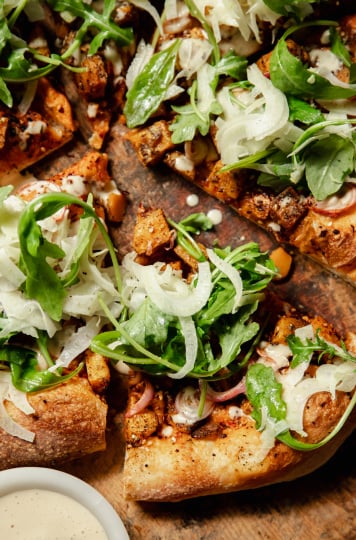 An up close, overhead shot of a round, vegan butternut squash flatbread topped with an arugula fennel salad and drizzles of a creamy white sauce.