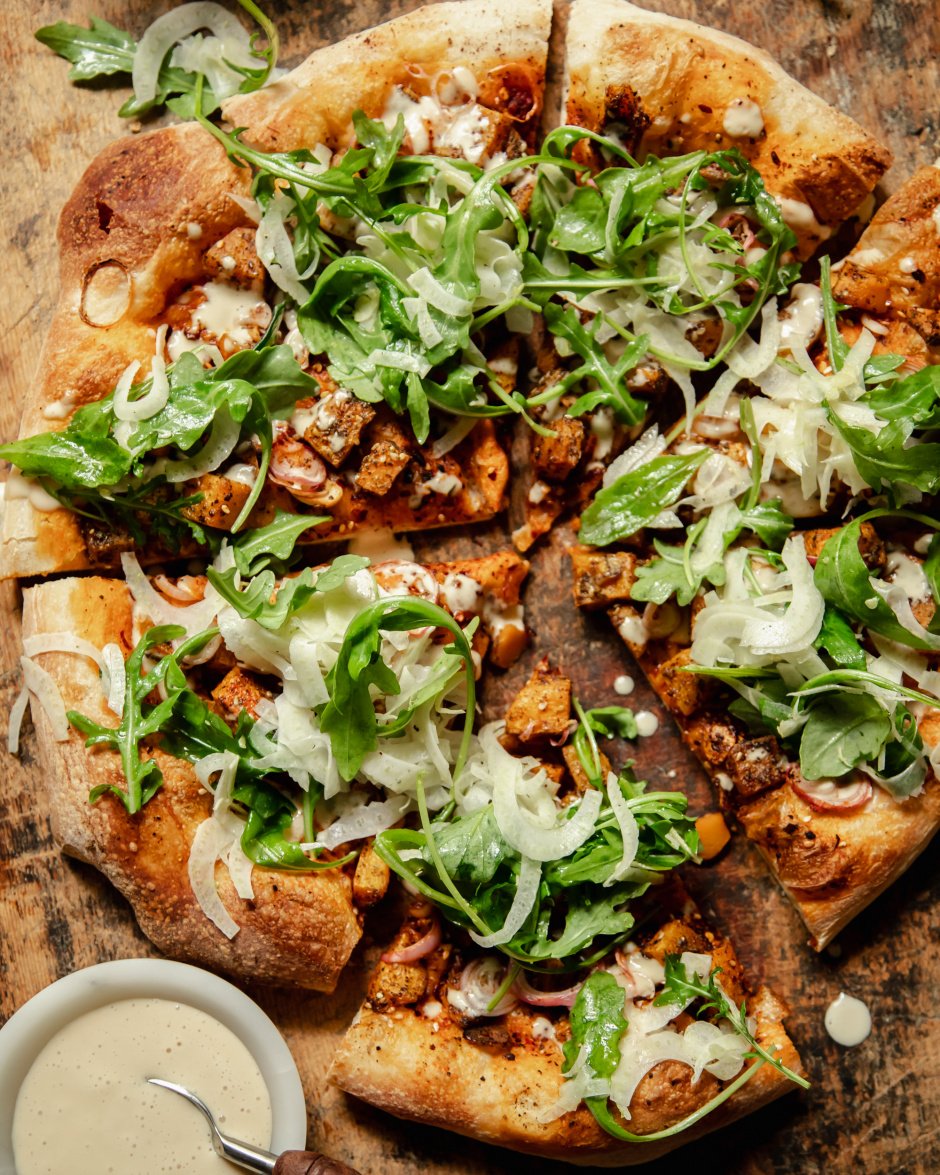 An overhead shot of a round, vegan butternut squash flatbread topped with an arugula fennel salad and drizzles of a creamy white sauce.
