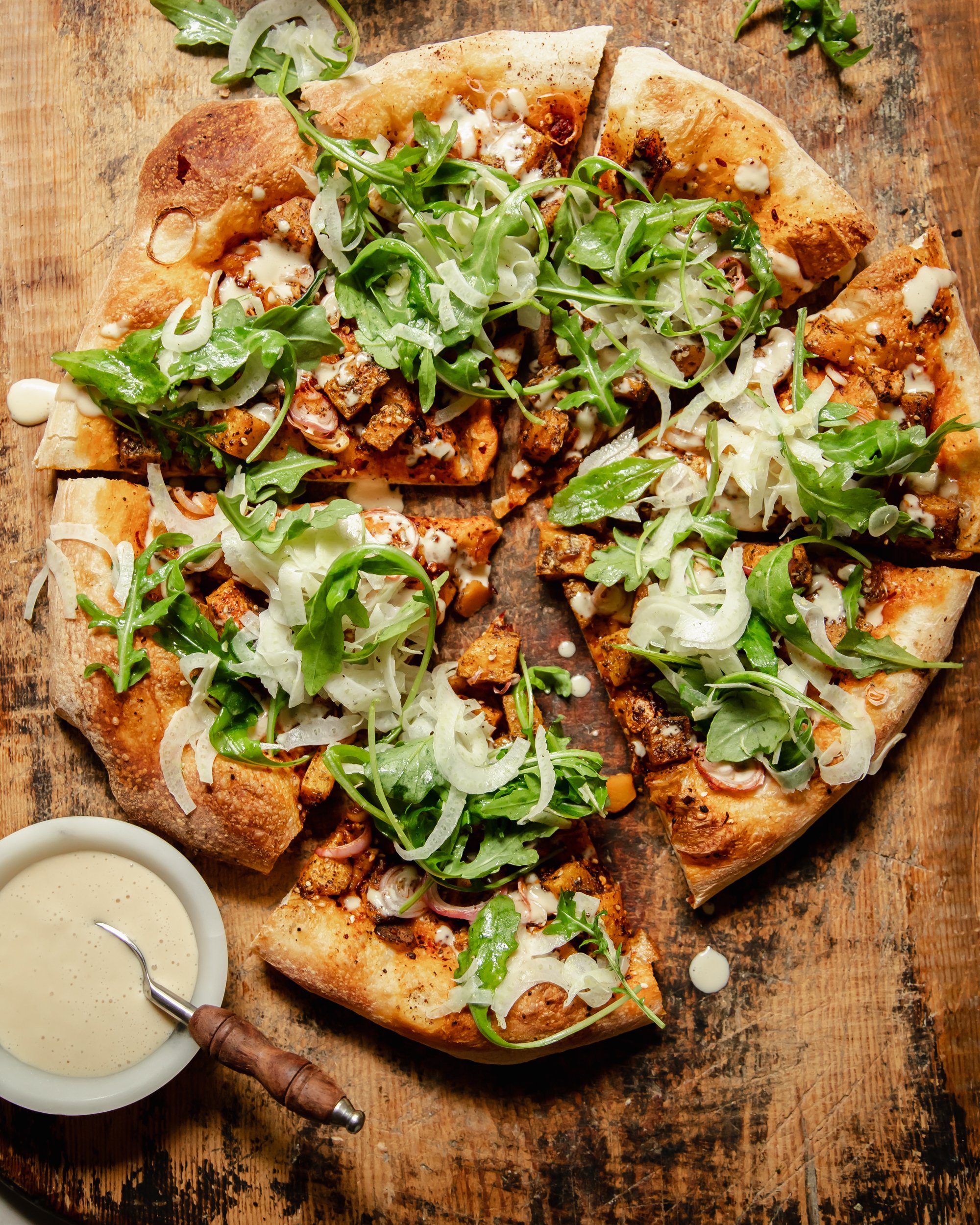An overhead shot of a round, vegan butternut squash flatbread topped with an arugula fennel salad and drizzles of a creamy white sauce.
